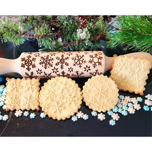 May include: A wooden rolling pin with a snowflake design is shown with four cookies that have been rolled with the pin. The cookies are on a black surface with white and blue sprinkles.