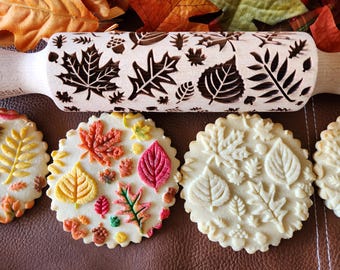 May include: A wooden rolling pin with a carved design of various autumn leaves. Three cookies are decorated with the same leaf design, showing the rolling pin's use.