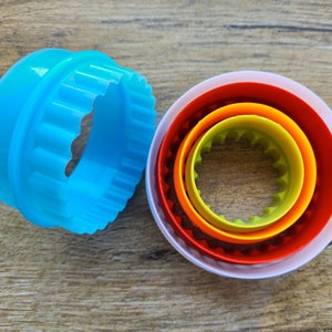 Puede incluir: Un juego de cinco cortadores de galletas redondos. El cortador más grande es blanco con un anillo rojo, naranja, amarillo y verde. El cortador más pequeño es azul claro con un borde festoneado.