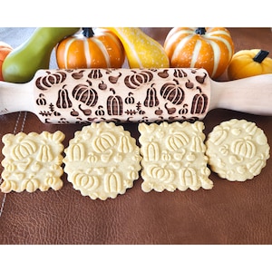 May include: A wooden rolling pin with a carved pumpkin design. Four cookies with the same pumpkin design are on a brown surface.