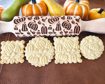 May include: A wooden rolling pin with a carved pumpkin design. Four cookies with the same pumpkin design are on a brown surface.