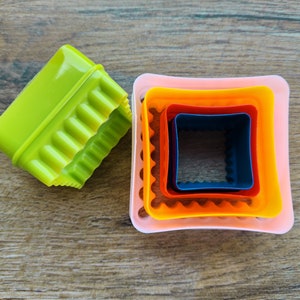 May include: Set of four square cookie cutters, nested inside each other. The cutters are pink, orange, red, and blue. The largest cutter has a scalloped edge.