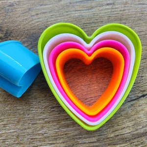 May include: Set of four heart-shaped cookie cutters in shades of green, white, pink, and orange. The cutters are nested inside each other, with the largest cutter on the outside and the smallest cutter on the inside.