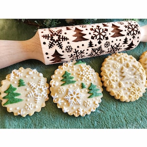Puede incluir: Un rodillo de madera con un diseño de árbol de Navidad y copo de nieve. El rodillo se utiliza para crear patrones decorativos en las galletas. Se muestran tres galletas con árboles de Navidad verdes y copos de nieve blancos.