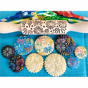 May include: A wooden rolling pin with a carved design of fireworks. The rolling pin is surrounded by cookies decorated with colorful firework designs.