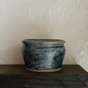 May include: A handcrafted ceramic pot with a dark teal glaze. The pot has a rounded shape with a slightly flared rim and a natural, unglazed base. The exterior features a speckled texture, adding visual interest to the piece. This decorative pot is suitable for plants or as a decorative item.