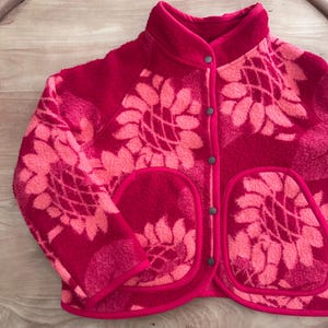 Peut inclure: Une veste en polaire à manches longues avec un motif de tournesols rose vif et rose clair. La veste présente un col montant, une fermeture boutonnée à l'avant avec des boutons-pression gris foncé et deux grandes poches plaquées avec bordure rose.