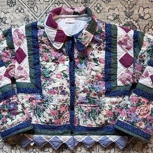 May include: A white quilted jacket with a floral print and patchwork design. The jacket has a button closure and a collar. The fabric is a mix of white, pink, purple, and green.