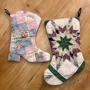 May include: Two handmade Christmas stockings. One is shaped like a boot with pink, blue, and floral patchwork. The other is a traditional stocking with a star pattern in red, green, and blue on a white background.