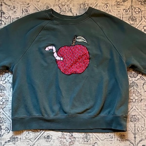 May include: A dark green sweatshirt with a crew neck and long sleeves. It has an embroidered red apple with a white worm and a green leaf on the front. The sweatshirt is laid flat on a patterned surface.