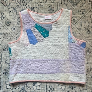 Peut inclure: Un gilet matelassé avec un motif patchwork. Le gilet présente une variété de couleurs pastel, dont le bleu clair, le lavande et le sarcelle, avec une bordure rose. Le gilet a un motif en losange et le texte "Trible Edit" sur l'étiquette.