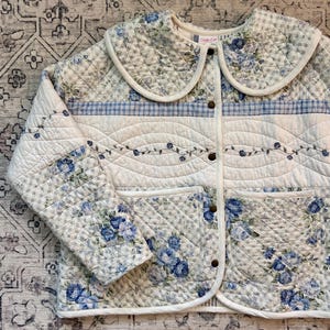 May include: A quilted jacket featuring a blue and white floral pattern mixed with a blue and white gingham check. The garment has a large rounded collar, long sleeves, two front patch pockets, and a front button closure with metal snaps.