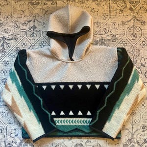May include: A cream fleece pullover with a hood and a black neck gaiter. The design features geometric patterns in black, teal, and tan. The bottom hem and sleeve cuffs are trimmed with black fabric.
