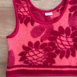 May include: A sleeveless top made of textured terry cloth fabric in shades of bright pink and deep magenta. The garment features a large floral pattern, a round neckline, and solid pink trim along the edges. A white label reads Textile Edit.