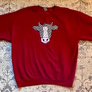 May include: A red sweatshirt featuring a cow face design. The cow's head is white and pink with a black and white checkered pattern. The sweatshirt has long sleeves and a crew neck.