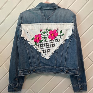 May include: A blue denim jacket with a white crocheted patch on the back. The patch features pink flowers and green leaves.