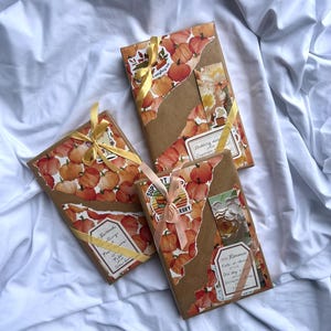 Puede incluir: Tres regalos envueltos con papel marrón y papel decorativo con temática de calabazas. Cada regalo está adornado con una cinta y pegatinas. Los regalos están colocados sobre un fondo de tela blanca.