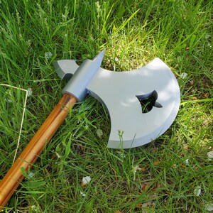 May include: A silver and gray battle axe with a wooden handle lies on a bed of green grass. The axe head has a decorative cutout. The handle is a light brown color.
