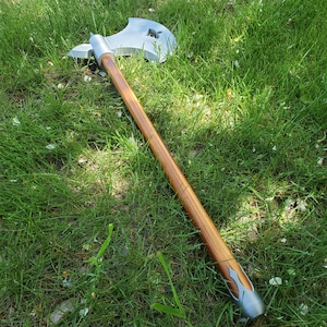 May include: A silver and wood axe with a long wooden handle. The axe head has a unique shape with a star-shaped cutout. The handle is a light brown color, and the axe is resting on green grass.