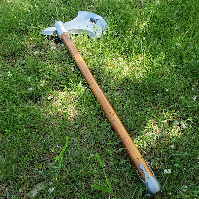 May include: A silver and wood axe with a long wooden handle. The axe head has a unique shape with a star-shaped cutout. The handle is a light brown color, and the axe is resting on green grass.