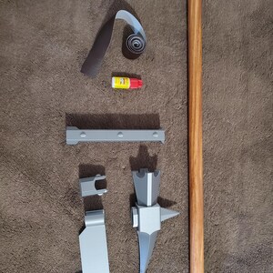 May include: An assortment of craft supplies and components, including a wooden dowel, gray metal pieces, sandpaper, and a small tube of glue. The items are arranged on a brown surface, suggesting a DIY project or craft kit.