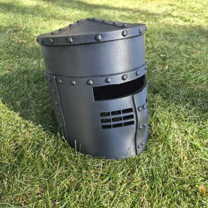 Peut inclure: Un heaume gris foncé à sommet plat, doté d'une fente oculaire rectangulaire horizontale et d'une série de petits trous de ventilation rectangulaires sur la face avant. Le casque est orné d'une série de rivets en relief sur les bords.