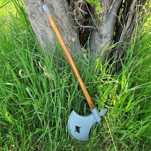May include: Axe with a light grey head and a wooden handle. The axe head has a decorative cutout. The handle is brown with a grey top. The axe is leaning against a tree trunk in a grassy area.