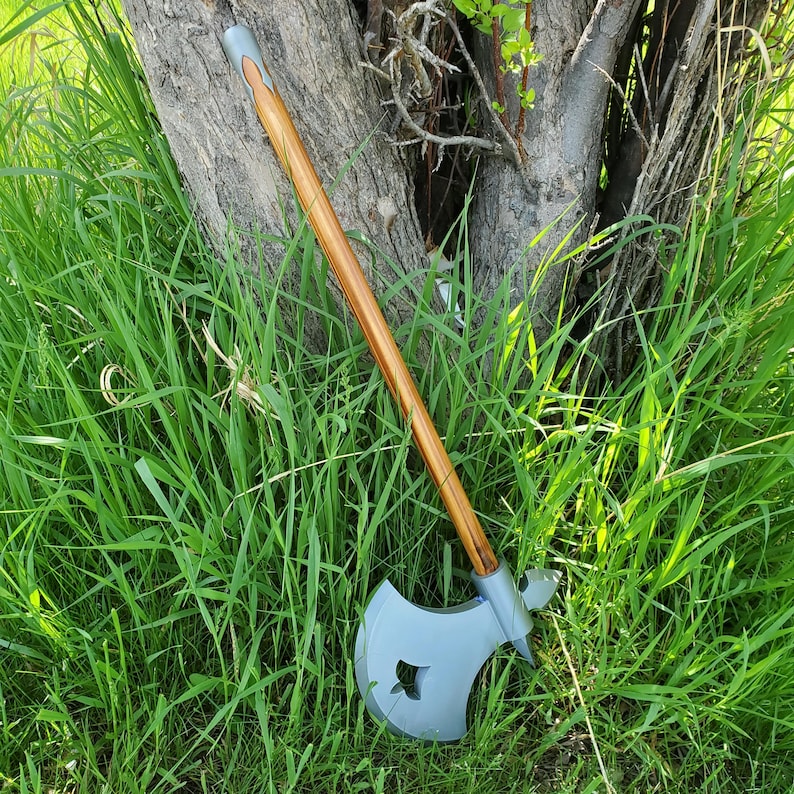 May include: Axe with a light gray head and a wooden handle. The axe head has a decorative cutout. The handle is brown with a gray top. The axe is leaning against a tree trunk in a grassy area.