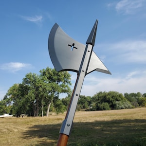 May include: A large, gray battle axe with a cross-shaped cutout in the blade. The axe has a long wooden handle and is set against a backdrop of a blue sky and grassy field.