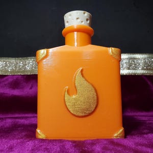May include: Orange, square-shaped bottle with a gold flame design on the front. The bottle has gold accents on the corners and a white and tan stopper. The bottle is sitting on a purple surface.