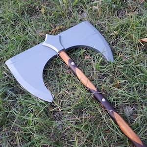 May include: A large, double-bladed axe with a gray head and a wooden handle. The handle is a combination of light and dark brown wood, with dark brown accents and silver rivets. The axe is lying on a bed of green grass.