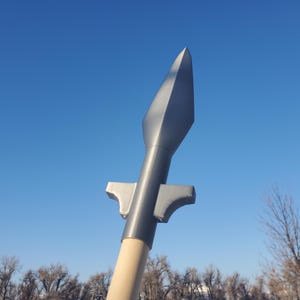 Può includere: Una lancia con una punta di freccia in metallo grigio e un'asta di legno. La punta della lancia ha una punta affilata ed è montata su un'asta cilindrica. L'asta è in legno di colore chiaro. Lo sfondo è un cielo azzurro.
