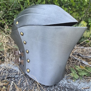 Op de afbeelding: Een metallic grijze ridderhelm met een glad, gebogen vizier en een puntig voorstuk. De zijkant is bevestigd met een verticale rij van zes messingkleurige klinknagels en twee bruine leren riemen met zilverkleurige metalen gespen voor verstelling.