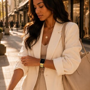 Puede incluir: Un blazer de color crema sobre una parte superior de canalé beige, combinado con un bolso tote beige. Un reloj inteligente con una correa de rayas amarillas y azules se lleva en la muñeca. El conjunto incluye un collar con colgante y aros dorados.
