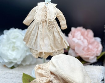 Könnte beinhalten: Ein weißes Puppenkleid mit Spitzenbesatz und ein passender weißer Hut mit einer Schleife aus Spitze. Das Kleid und der Hut werden auf einer weißen Puppe und einer weißen Oberfläche präsentiert.