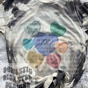 May include: A white t-shirt with a black tie-dye pattern. The shirt features a design of colorful candy hearts with text that reads "Will pcs with you", "Bad girl", "Dependa", "Raw", "I had at Trader", "Be my sponsor", and "Upscale cutie".