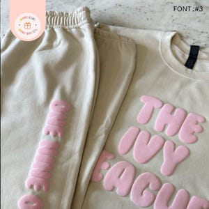 May include: A beige sweatsuit set featuring a long-sleeved shirt and sweatpants. The shirt displays the text "THE IVY LEAGUE" in pink, puffy letters. The sweatpants have the text "ONE MORE" in pink, puffy letters. The set is from "MORE LOVE MORE LIFE CO."