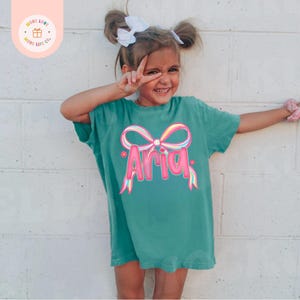 May include: Teal t-shirt with the name "Aria" in pink bubble letters, adorned with a colorful bow graphic. The shirt is worn by a young girl with a white bow in her hair. The shirt has a relaxed fit.