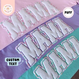 K&ouml;nnte beinhalten: Nahaufnahme von drei T-Shirts in Rosa, Lila und T&uuml;rkis. Jedes Shirt zeigt das Wort "MAMA" in wei&szlig;en, gepufften, gestickten Buchstaben mit einem farbenfrohen Rand. Die Shirts sind von der Marke "MORE LOVE MORE LIFE CO." und bieten individuelle Textoptionen.