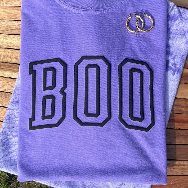 May include: Purple t-shirt with the word "BOO" printed in black block letters.