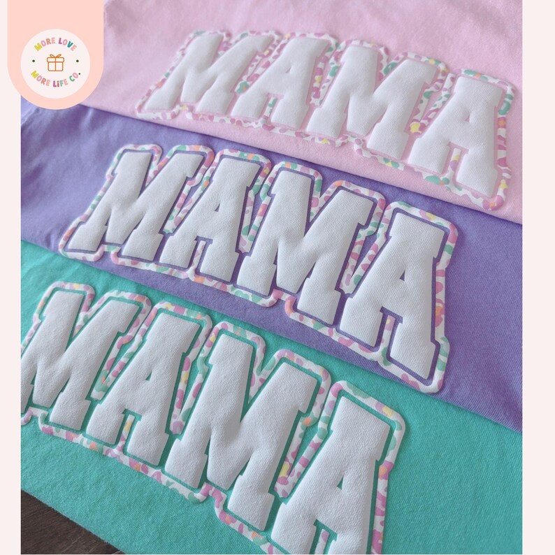 K&ouml;nnte beinhalten: Nahaufnahme von drei pastellfarbenen T-Shirts: rosa, lila und t&uuml;rkis. Jedes Shirt zeigt das Wort "MAMA" in gro&szlig;en, wei&szlig;en, gepufften Buchstaben mit einem bunten, gemusterten Rand. Die Shirts sind ordentlich gestapelt und zeigen das Design.