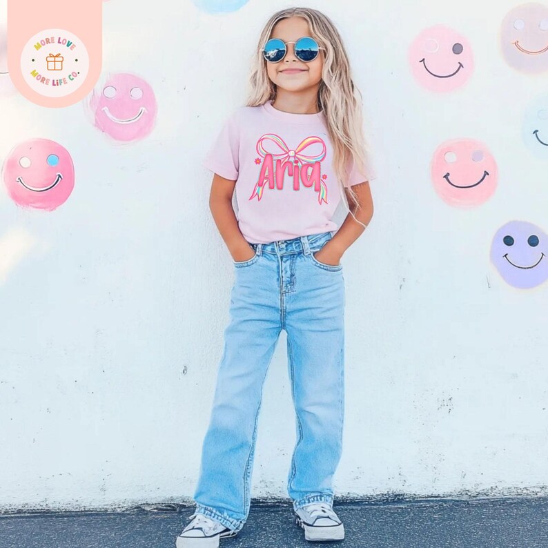 May include: A young girl wearing a light pink t-shirt with the name "Aria" in a bow design, paired with light blue jeans and white sneakers. She is wearing sunglasses. Smiley face decorations are on the white wall behind her.