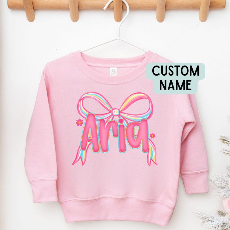 May include: A pink sweatshirt with the name "Aria" in a bubble font, accented with a rainbow bow and flowers. The words "CUSTOM NAME" are in a light blue box. The sweatshirt is hanging on a white hanger.