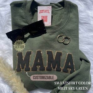 May include: A green sweatshirt with the word "MAMA" in black puff lettering with gold glitter outlining. The sweatshirt is customizable and the color is military green.