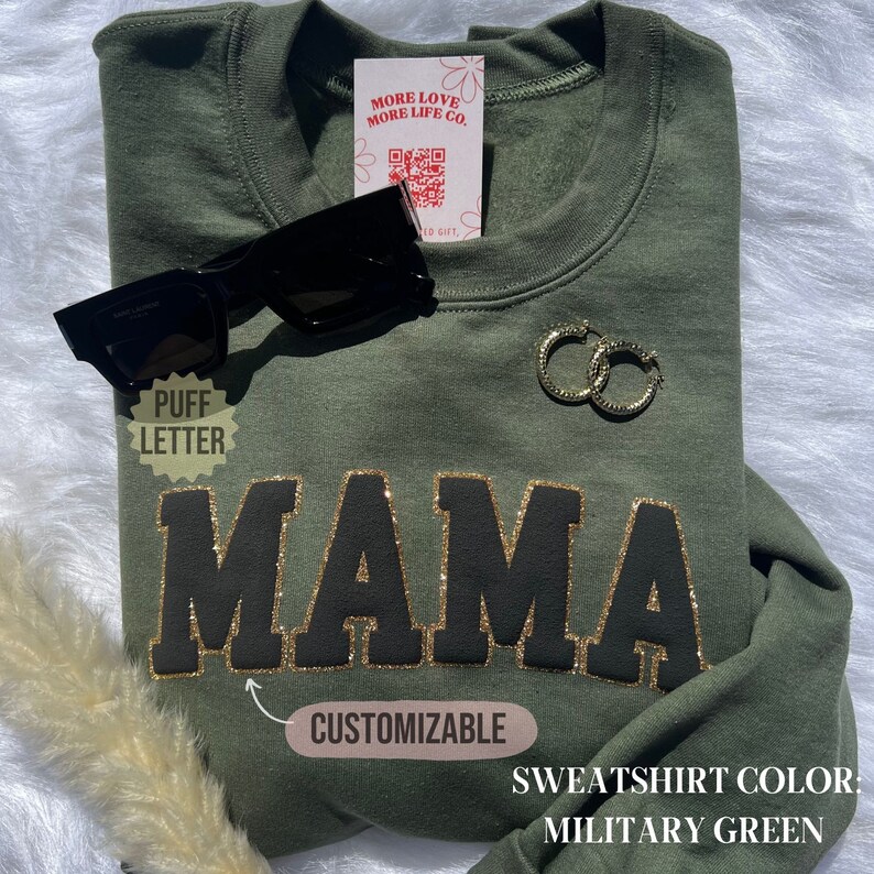 May include: A green sweatshirt with the word "MAMA" in black puff lettering with gold glitter outlining. The sweatshirt is customizable and the color is military green.