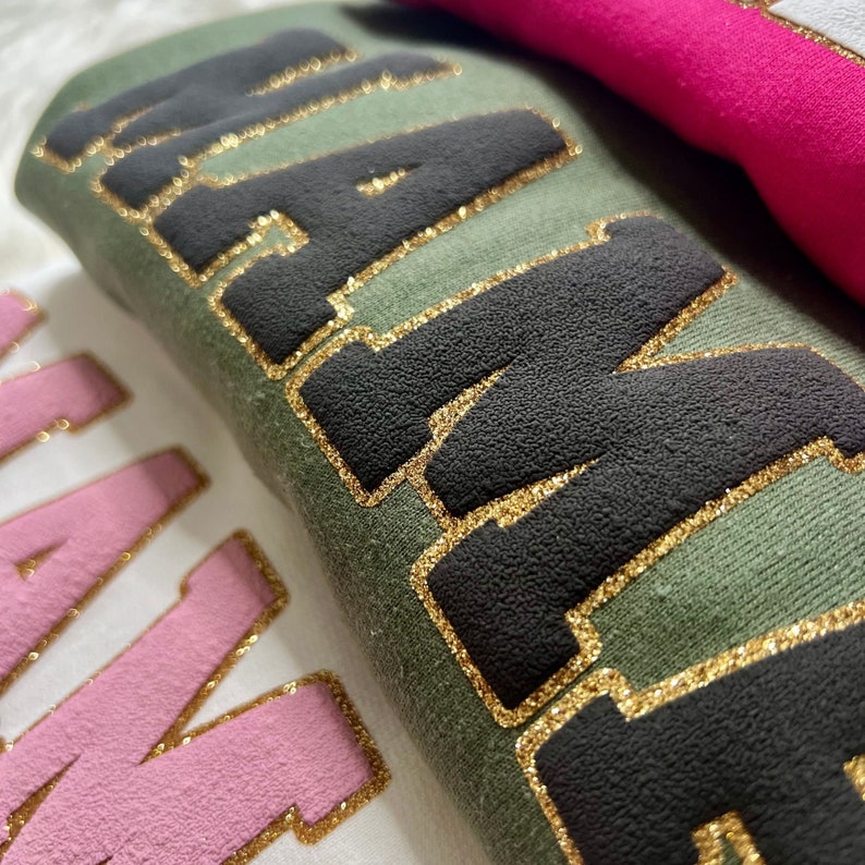 May include: Close-up of two shirts with the word "Mama" written in gold glitter on each. The shirt on top is green with black lettering, and the shirt on the bottom is white with pink lettering.
