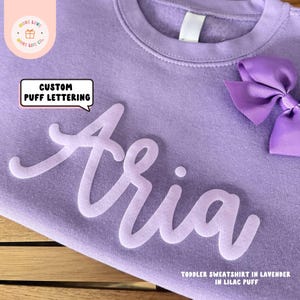 May include: A lavender toddler sweatshirt with the name "Aria" in white puffy lettering. The sweatshirt has a crew neck and is made of soft, comfortable fabric.