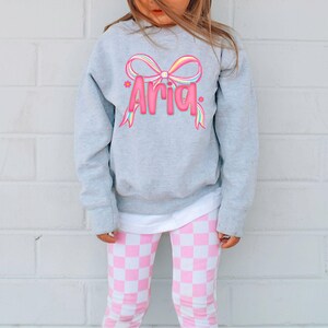 May include: A gray sweatshirt with the name "Aria" in pink, with a colorful bow design. The child is wearing pink and white checkered leggings. The outfit is casual and suitable for everyday wear.