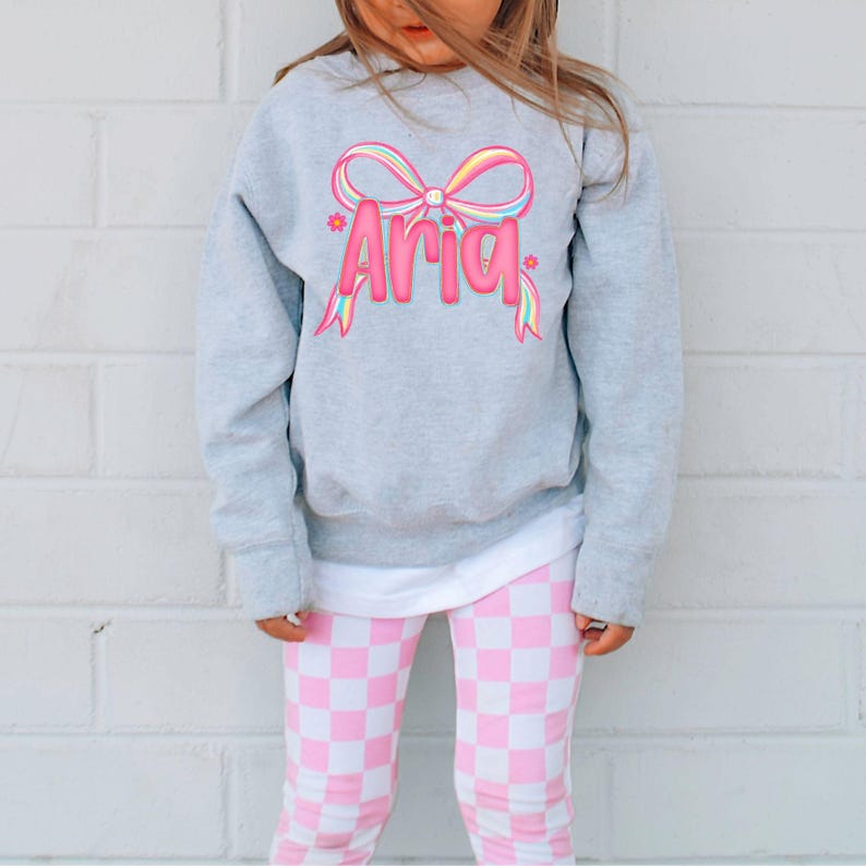 May include: A gray sweatshirt with the name "Aria" in pink, with a colorful bow design. The child is wearing pink and white checkered leggings. The outfit is casual and suitable for everyday wear.