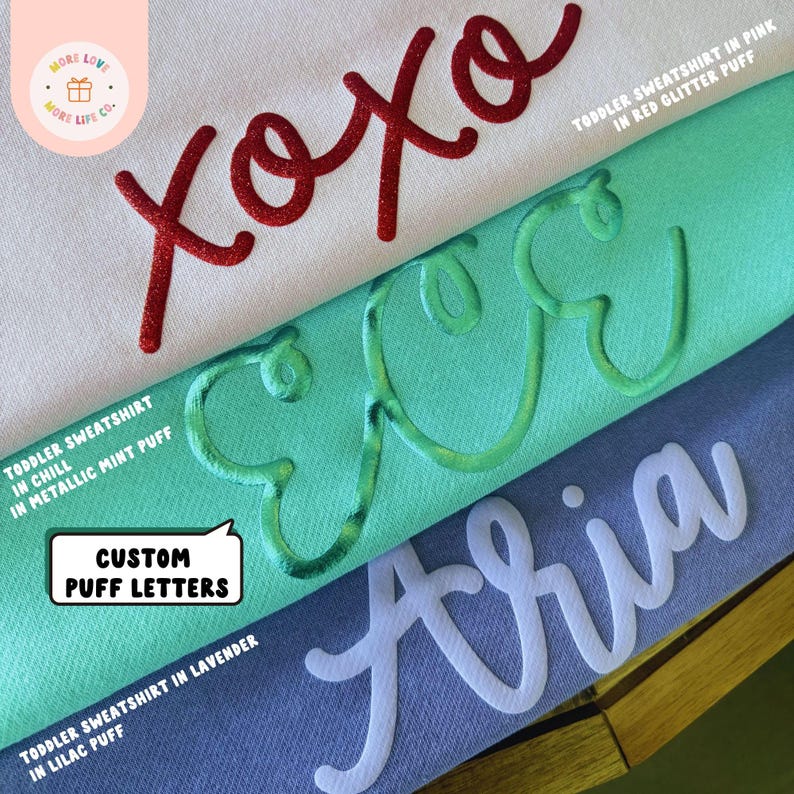 May include: Three toddler sweatshirts with custom puff lettering. The top sweatshirt is white with red glitter puff lettering that says "xoxo". The middle sweatshirt is turquoise with metallic mint puff lettering that says "xoxo". The bottom sweatshirt is lavender with lilac puff lettering that says "Aria".